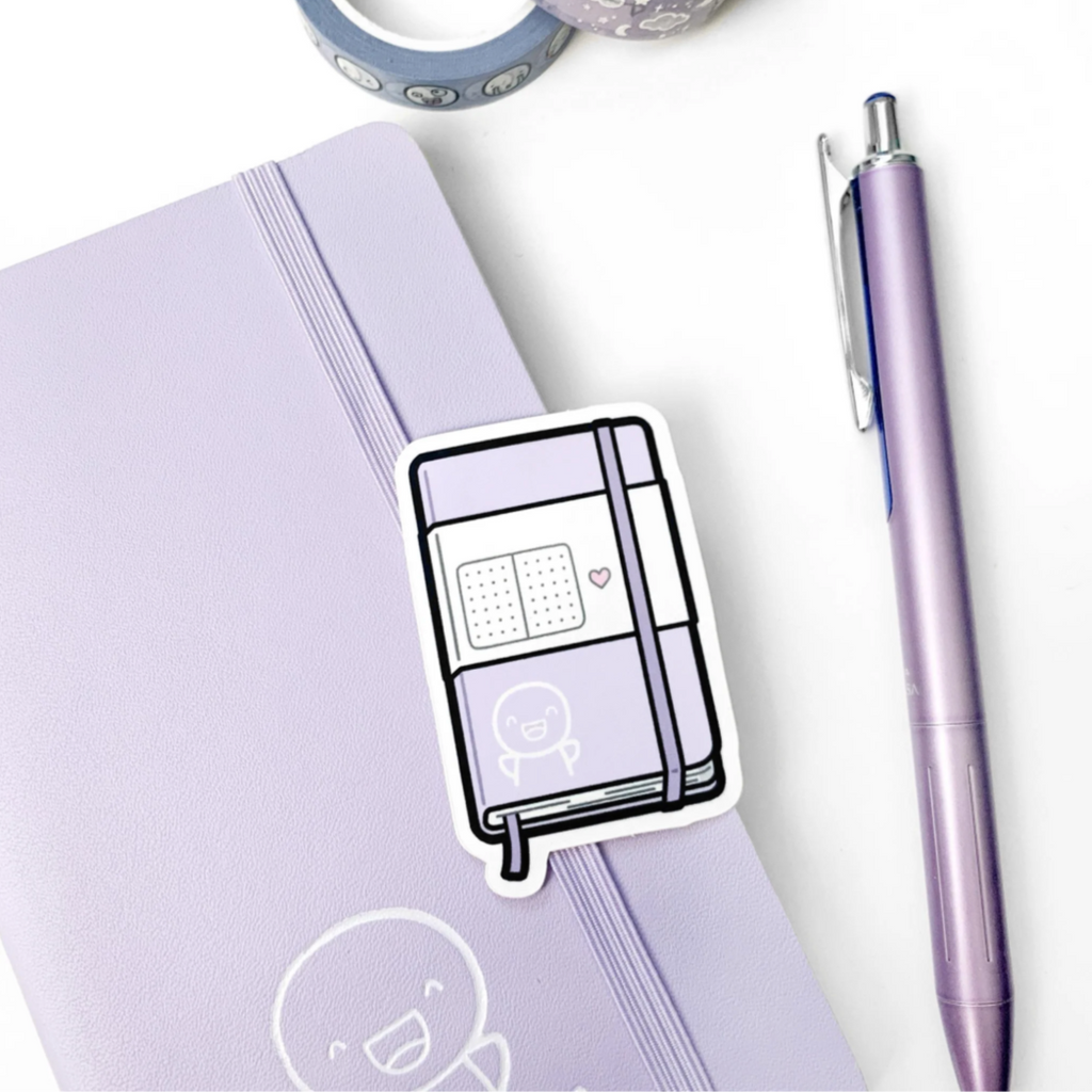 Sticker of a cartoonish book on a purple notebook with a pen and tape in the background