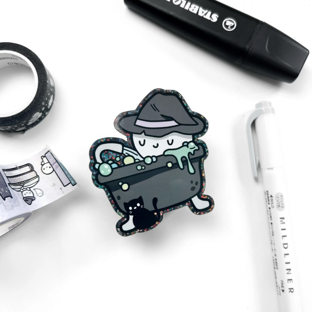 Cartoon character in a witch hat sitting in a cauldron with stationery items on a white background